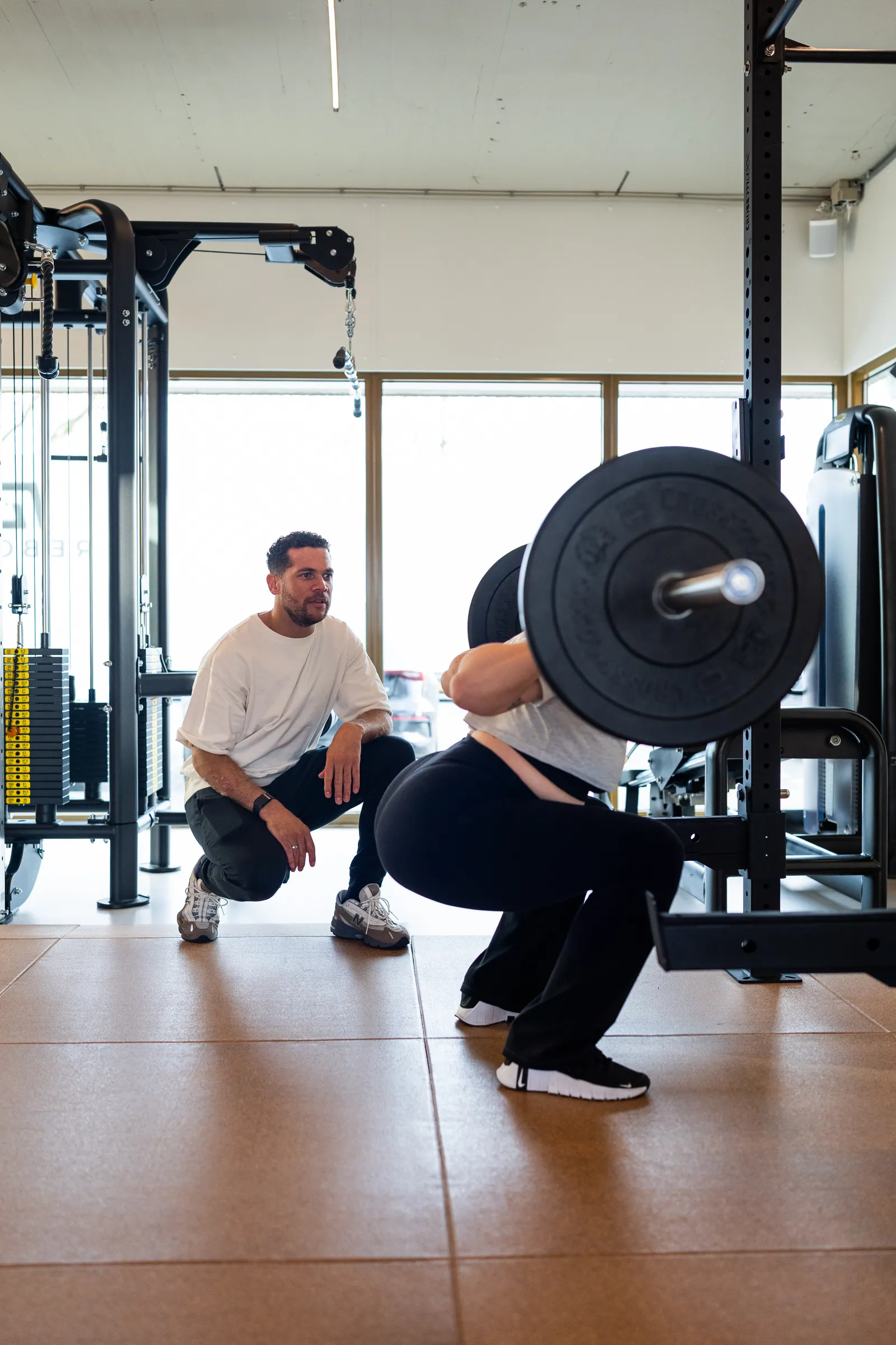 Coach knielt naast atlete die squat doet met barbell bij ramen