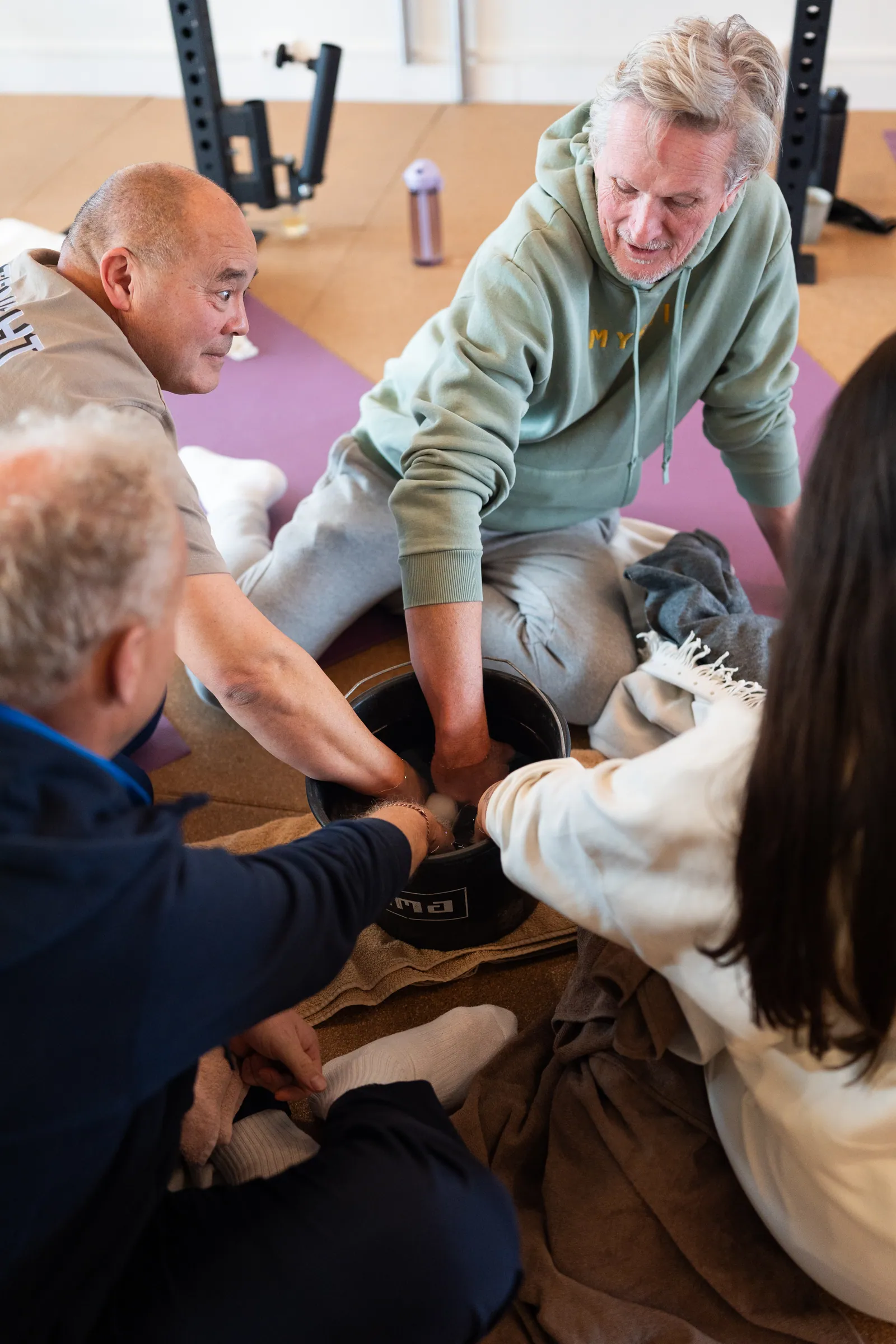 Groep leunt naar centrale ijsemmer op mat tussen krachttrainingrek en vloer.