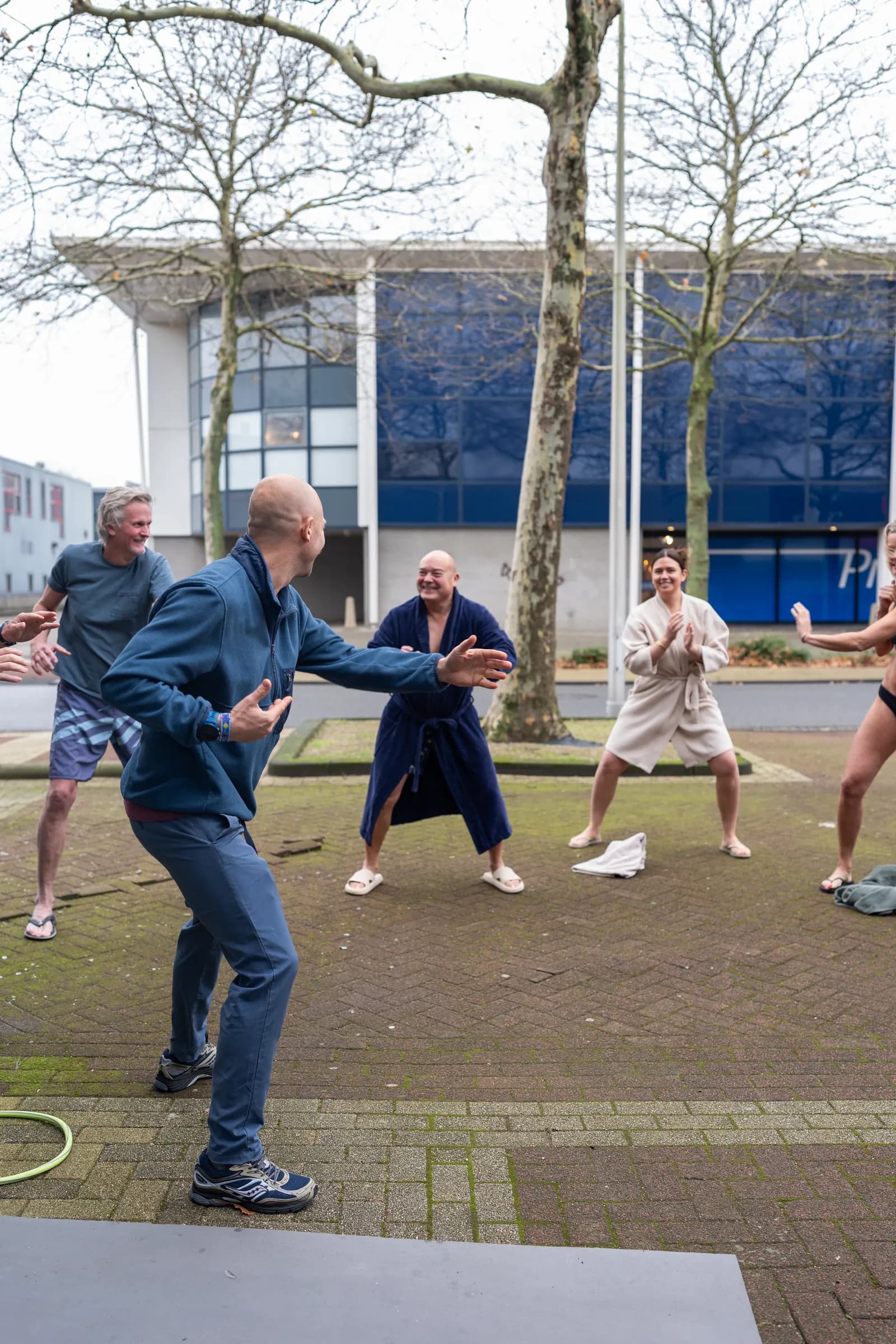 Coach in blauwe fleece leidt groep in badjassen tijdens bewegingsoefening buiten.