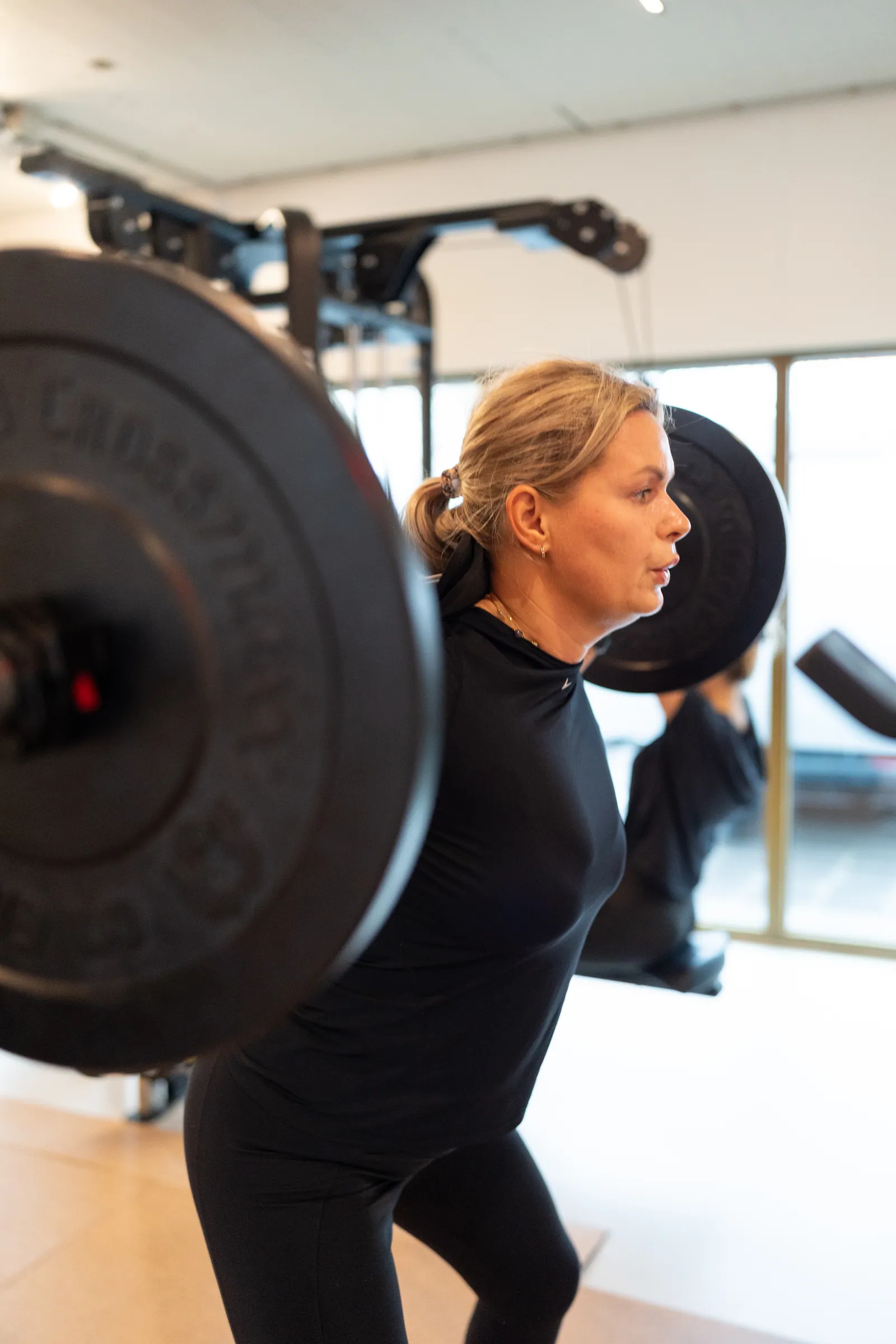 Vrouw in back squat met barbell; wazige schijf op voorgrond, ramen achter.