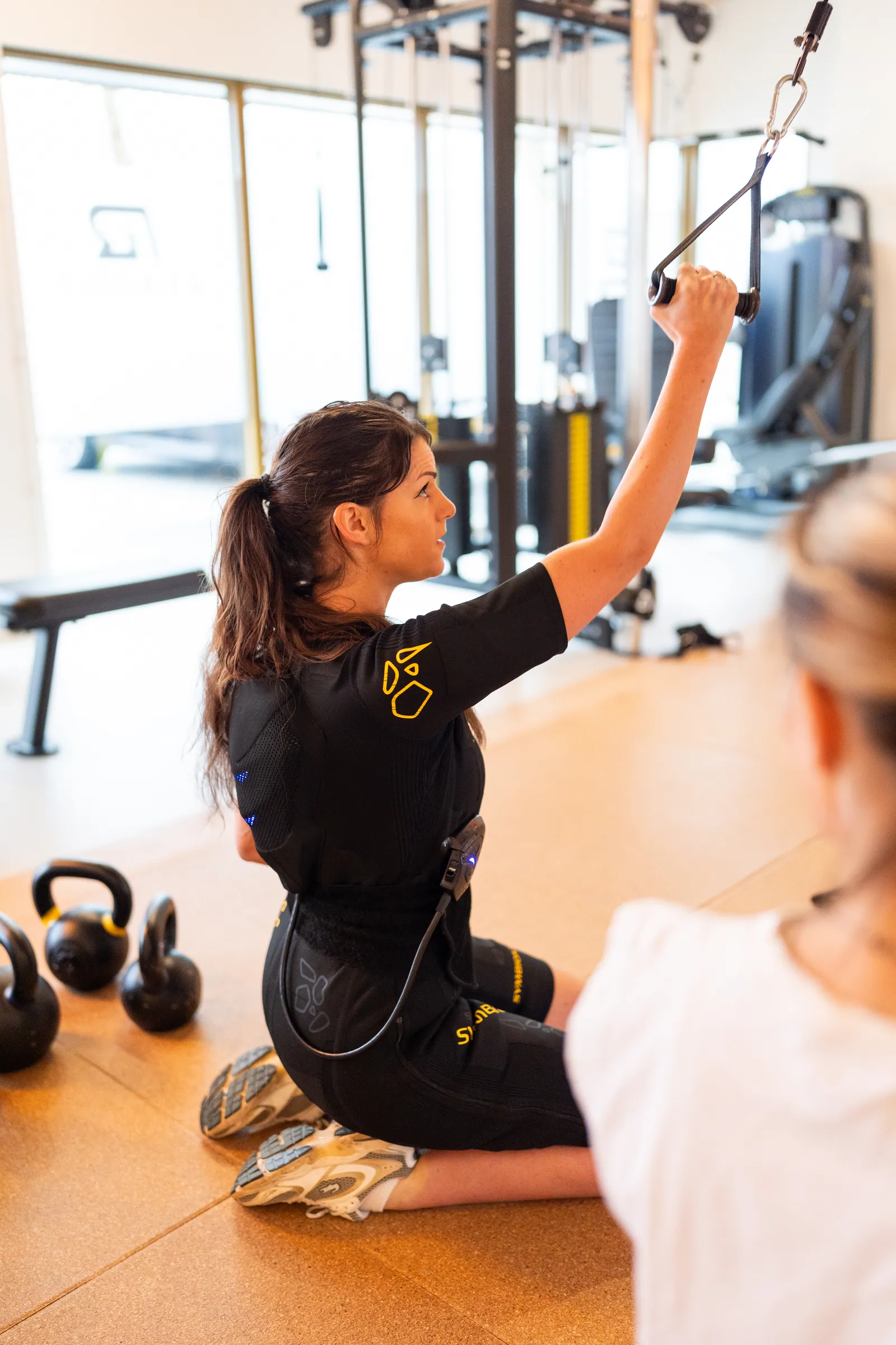 Vrouw in EMS-pak knielt en trekt aan een kabel; kettlebells op de kurkvloer.